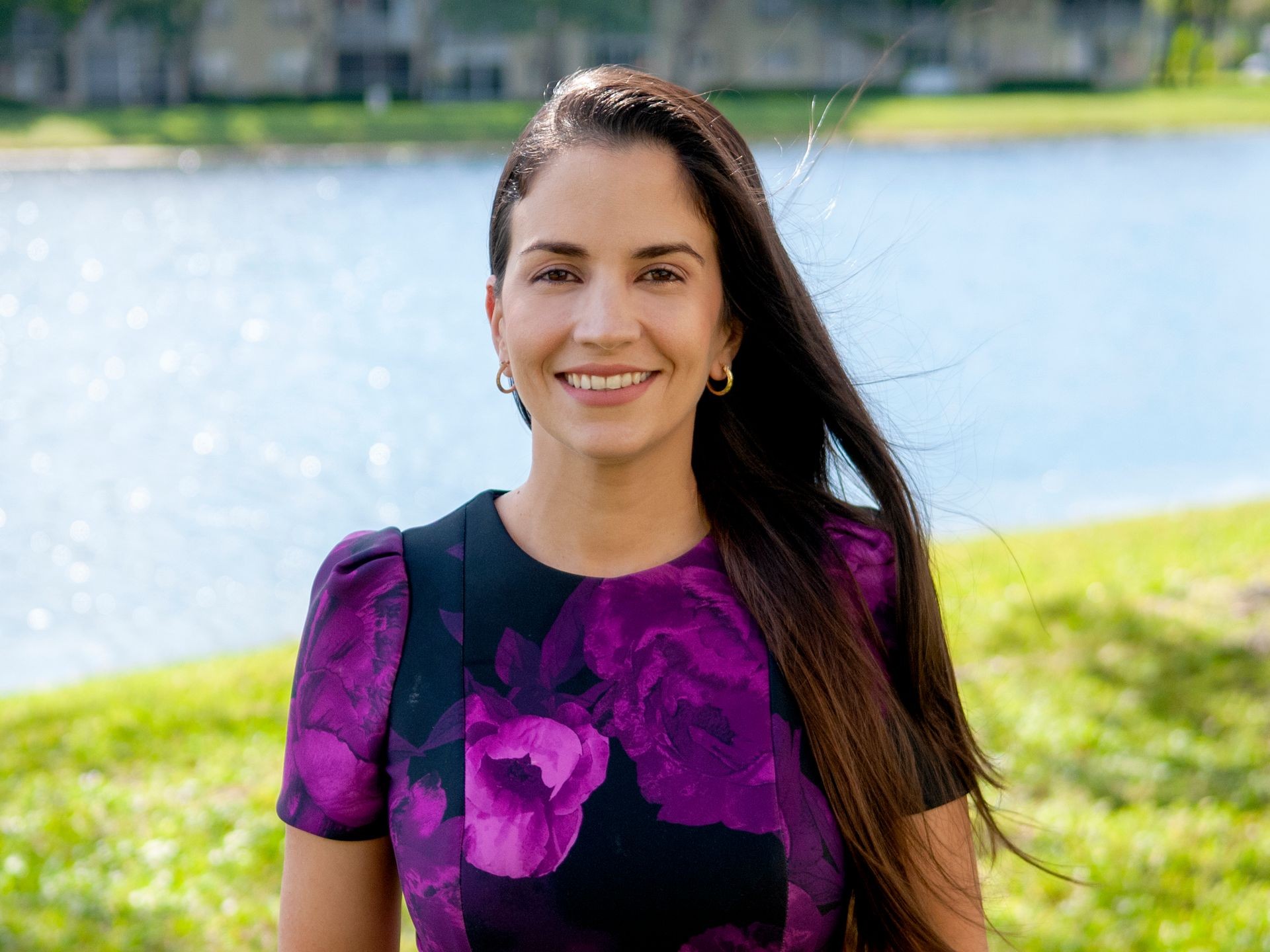 Smiling woman in a purple floral dress standing by a lake on a sunny day.