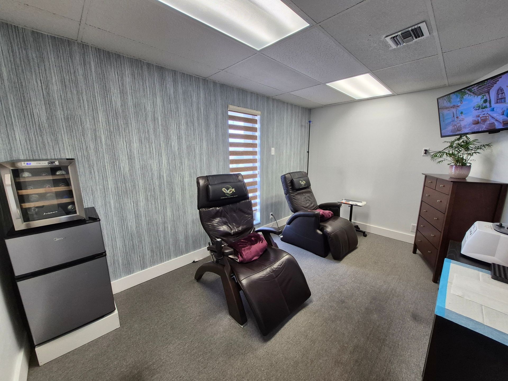 Comfortable seating area with massage chairs, small fridge, and TV in a modern therapy room.