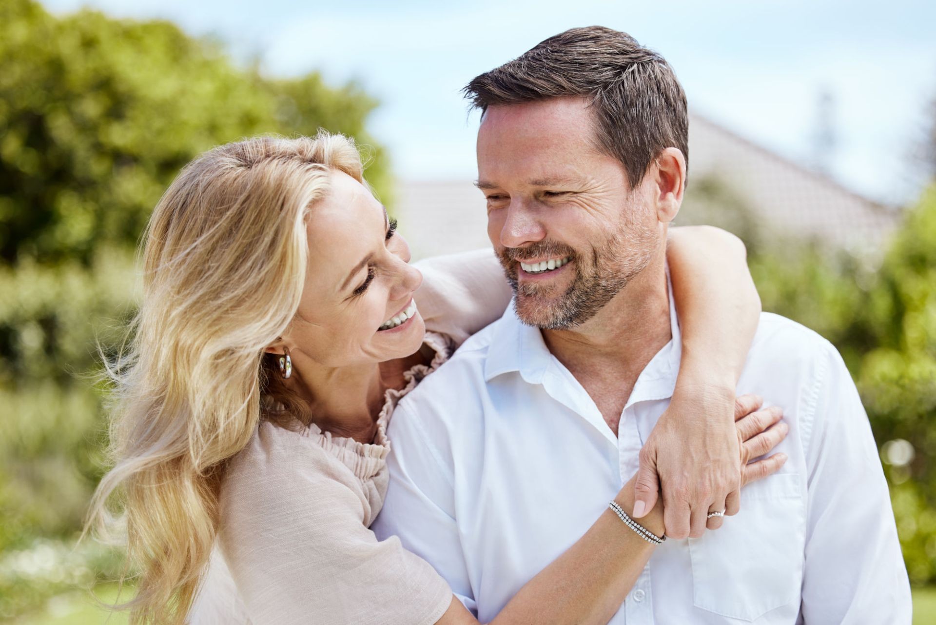 Smiling couple embracing outdoors in a garden setting, with trees and sky in the background.