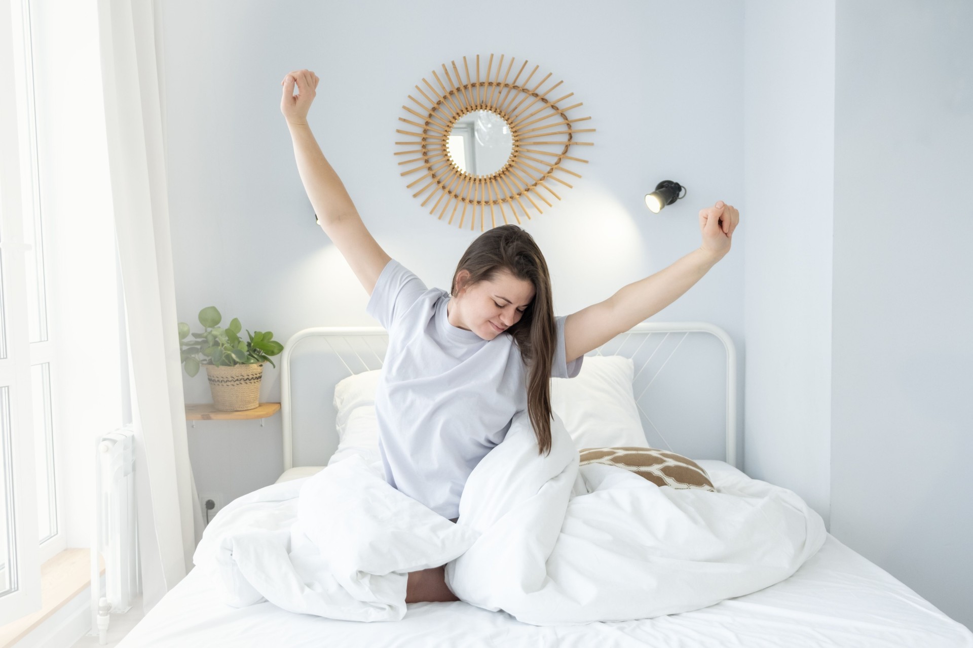 Happy cheerful girl sitting on bed after good sleep. Young woman enjoying morning on bed at home